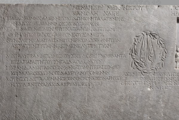 Family tombstone with epitaph, unknown workshop, Greek inscriptions, restitution, sepulchral funerary architecture, riders iconographic, wreaths, laurel wreaths, ribbon ornament, horses iconographic, sepulchral sculpture artist, rectangular marble slab, diagonally cracked. The block bears an inscription divided into three parts and two reliefs — a laurel wreath with a ribbon and a metope with a depiction of a young man on horseback accompanied by a slave . Both faces are damaged — perhaps hammered. The rider is dressed in a chiton and chlamys. The slave is exaggeratedly reduced in size in relation to the rider. He is dressed in a short girded chiton and holds a spear in his hand. 1st century BC–1st century AD, Cyzicus, Asia Minor, Greek inscription, grave inscription, stele, gravestone, bas-relief. Height 76 cm, width 203 cm, depth 11 cm, weight 450 kg., A stone slab with a rough, light gray surface carved with a grid of inscribed lines occupying the left and center areas and a carved ...