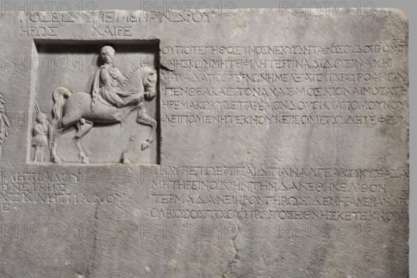 Family tombstone with epitaph, unknown workshop, Greek inscriptions, restitution Braniewo; Lyc.Hosianum, sepulchral funerary architecture, riders iconographic, wreaths, laurel wreaths, ribbon ornament, horses iconographic, sepulchral sculpture artist, rectangular marble slab, diagonally cracked. The block bears an inscription divided into three parts and two reliefs — a laurel wreath with a ribbon and a metope with a depiction of a young man on horseback accompanied by a slave . Both faces are damaged — perhaps hammered. The rider is dressed in a chiton and chlamys. The slave is exaggeratedly reduced in size in relation to the rider. He is dressed in a short girded chiton and holds a spear in his hand. 1st century BC–1st century AD, Cyzicus, Asia Minor, Greek inscription, grave inscription, stele, gravestone, bas-relief; height 76 cm, width 203 cm, depth 11 cm, weight 450 kg, A carved stone slab in pale gray with a recessed rectangular panel showing a relief of a person seated on a hor...
