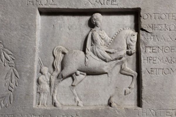 Family tombstone with epitaph, unknown workshop, Greek inscriptions, restitution Braniewo; Lyc. Hosianum, sepulchral funerary architecture, riders iconographic, wreaths, laurel wreaths, ribbon ornament, horses iconographic, sepulchral sculpture artist, rectangular marble slab, diagonally cracked. The block bears an inscription divided into three parts and two reliefs—a laurel wreath with a ribbon and a metope with a depiction of a young man on horseback accompanied by a slave . Both faces are damaged—perhaps hammered. The rider is dressed in a chiton and chlamys. The slave is exaggeratedly reduced in size in relation to the rider. He is dressed in a short girded chiton and holds a spear in his hand. 1st century BC–1st century AD, Cyzicus, Asia Minor, Greek inscription, grave inscription, stele, gravestone, bas-relief; height 76 cm, width 203 cm, depth 11 cm, weight 450 kg., A carved stone relief shows a raised rectangular panel with a sculpted scene of a person seated on a horse and a ...