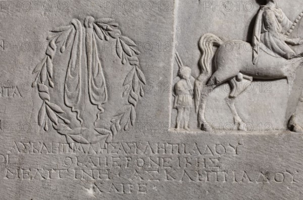 Family tombstone with epitaph, unknown workshop, Greek inscriptions, restitution Braniewo ; Lyc.Hosianum, sepulchral funerary architecture, riders iconographic, wreaths, laurel wreaths, ribbon ornament, horses iconographic, sepulchral sculpture, rectangular marble slab, diagonally cracked. The block bears an inscription divided into three parts and two reliefs - a laurel wreath with a ribbon and a metope with a depiction of a young man on horseback accompanied by a slave . Both faces are damaged - perhaps hammered. The rider is dressed in a chiton and chlamys. The slave is exaggeratedly reduced in size in relation to the rider. He is dressed in a short girded chiton and holds a spear in his hand. 1st century BC - 1st century AD, Cyzicus, Asia Minor, Greek inscription, grave inscription, stele, gravestone, bas-relief; height 76 cm, width 203 cm, depth 11 cm, weight 450 kg., A carved stone relief showing a circular laurel wreath with draped ribbons on the left and a partial scene on the ...