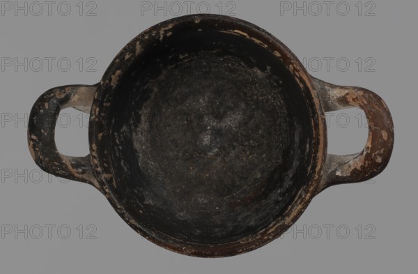 Miniature skyphos. Reddish clay, black-brown firnis poorly covering the outside on the foot, at the bottom of the body, the outer parts of the handles, the rim and the inside. The zone between the handles is white-ground, black-figure. Decoration: A - seated human figure in right profile; B - standing human figure facing right and quadruped facing left. 1st quarter of 5th century BC, Attica. Vessel, skyphos; height 4 cm, width 5.2 cm., unknown Attic workshop. Materials/technique: white-ground and black-figure pottery; miniature vessel; palmette ornament., A top-down view of a round, shallow bowl or pot with two opposing handles, showing a worn, rough interior and exterior surface; the object displays a mix of black, dark gray, brown, beige, and hints of off-white, with patches where the darker surface has chipped to reveal lighter underlying material, set against a plain gray background.