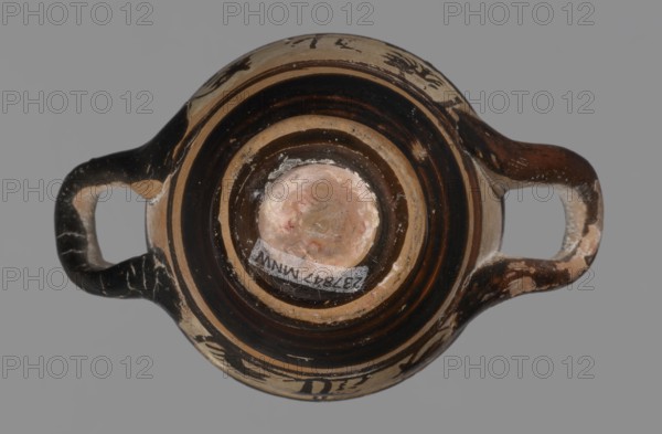 Miniature skyphos. Reddish clay, black-brown firnis poorly covering the outside on the foot, at the bottom of the body, the outer parts of the handles, the rim and the inside. The zone between the handles is white-ground, black-figure. Decoration: A - seated human figure in right profile; B - standing human figure facing right and quadruped facing left., Attic workshop; 1st quarter of 5th century BC, Attica Athens, Ancient Greece. Vessel skyphos, height 4 cm, width 5.2 cm. Palmettes ornament; miniature vessel; white-ground and black-figure pottery., A top-down view of a small two-handled vessel with a rounded body and a central circular base area, showing concentric bands of dark brown and beige on the interior and exterior surfaces; the handles extend horizontally on either side and show worn areas revealing lighter beige beneath a dark brown surface; the central base has a pale, slightly pinkish beige patch with some irregular discoloration and residue; the vessel surface shows chipp...