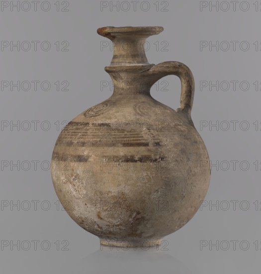 Jug with a flat foot, spherical body, high narrow neck with a strongly tilted spout and a small vertical handle supported by arms and a ring on the neck. Above the maximum diameter: a black band and a set of five parallel lines framed by thicker bands. Arms bear four sets of concentric circles arranged symmetrically on either side of the vessel's axis. Handle decorated with transverse lines. Painted pottery jug, Cypriot, Geometric Period III, 850–750 BC. Vessel; height 10.5 cm, width 7.8 cm., A rounded ceramic jug with a narrow neck, flared rim, and single handle, showing an overall weathered surface in shades of beige, tan, brown, and gray; the body displays a few darker brown horizontal bands and faint circular painted motifs near the shoulder, with areas of surface wear, patina, and mottled discoloration in lighter and darker beige and gray tones, set against a plain gray background.