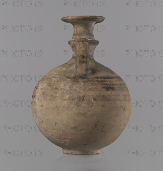 Cypriot jug, ca. 850–750 BC Cypriot Geometric Period III. Painted pottery jug with a flat foot, spherical body, high narrow neck with a strongly tilted spout, and a small vertical handle supported by arms and a ring on the neck. Decoration: above the maximum diameter a black band and a set of five parallel lines framed by thicker bands; on the arms four sets of concentric circles arranged symmetrically on the sides of the vessel's axis; handle decorated with transverse lines. Origin: Cyprus. Vessel jug. Height 10.5 cm, width 7.8 cm., A rounded clay vessel with a narrow neck and flared rim sits against a plain background, showing an aged surface with worn patches and chips; the body is bulbous with a short foot and a single handle rising from the shoulder to the neck. The visible colors include muted beige, tan, brown, gray, and hints of darker brown in the weathered bands and marks, with subtle variations of these tones across the surface.