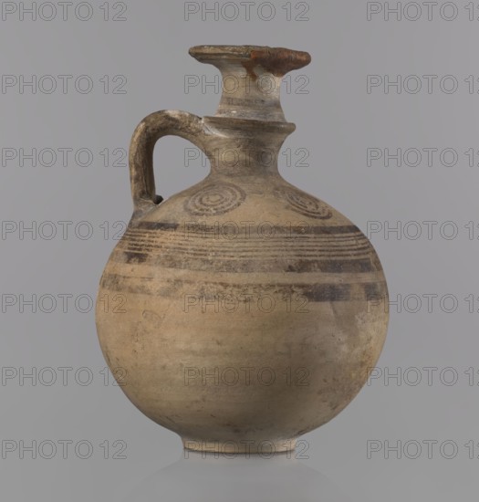 Cypriot jug, Jug with a flat foot, spherical body, high narrow neck with a strongly tilted spout, and a small vertical handle supported by arms and a ring on the neck. Above the maximum diameter: a black band and a set of five parallel lines framed by thicker bands. On the arms: four sets of concentric circles arranged symmetrically on the sides of the vessel's axis. Handle decorated with transverse lines. Painted pottery, Cypriot — Geometric Period III, 850–750 BC. Vessel jug. Height 10.5 cm; width 7.8 cm., A rounded ceramic jug with a narrow neck and a single curved handle, showing a worn surface with visible brushlike bands and circular motifs, in shades of beige, tan, brown, gray, and a few darker brown accents against a light gray background.