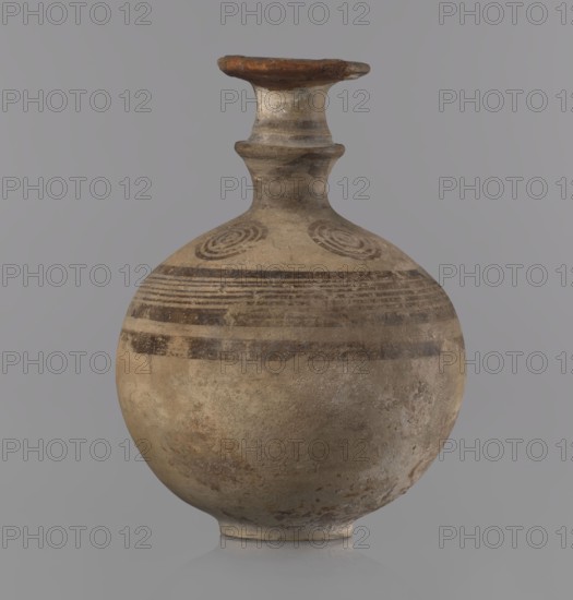 Cypriot jug, Jug with a flat foot, spherical body, high narrow neck with a strongly tilted spout, and a small vertical handle supported by arms and a ring on the neck. Above the maximum diameter: a black band and a set of five parallel lines framed by thicker bands. On the arms: four sets of concentric circles arranged symmetrically on the sides of the vessel's axis. Handle decorated with transverse lines. Painted pottery; vessel jug. Cypriot — Geometric Period III, 850–750 BC. Origin: Cyprus. Height 10.5 cm, width 7.8 cm., A rounded ceramic vessel with a narrow neck and flared rim sits against a neutral background; the vessel shows earthy tones of beige, tan, brown, and gray with darker brown painted bands and circular spiral motifs near the shoulder, a weathered surface texture with patches of lighter and darker discoloration, and a small flat base supporting its bulbous body.