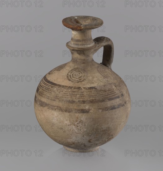 Jug, Cypriot, painted pottery, 850–750 BC Cypriot Geometric Period III. Jug with a flat foot, spherical body, high narrow neck with a strongly tilted spout, and a small vertical handle supported by arms and a ring on the neck. Decoration: above the maximum diameter a black band and a set of five parallel lines framed by thicker bands; the arms bear four sets of concentric circles arranged symmetrically on the sides of the vessel's axis; handle decorated with transverse lines. Medium: painted pottery. Function: vessel jug. Dimensions: height 10.5 cm, width 7.8 cm., A rounded ceramic vessel with a narrow neck and a rounded lip, featuring a single curved handle attached from the neck to the shoulder, resting on a small circular base; the surface shows a weathered, rough texture with areas of wear and pitting, and painted concentric circular motifs and horizontal banding around the shoulder; visible colors include beige, tan, brown, gray, and hints of darker brown in the painted decoration...