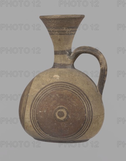 Cypriot jug, Cypriot pottery, Cypriot Bichrome Ware III. Jug on a low, straight foot. Globular body, flattened at the bottom. Quite narrow neck, funnel-shaped spout. Slightly flattened handle attached to the base of the neck and the upper part of the body. Painted decoration in stripes and linear patterns, in red-brown and black fading to brown. Near the spout is a wide red band, below it three black lines, the first adjacent to the red band. At the lower part of the neck, above the handle attachment, a narrow black band; another narrow black band at the base of the neck. The belly is decorated with three identical shooting target motifs on the front and sides: a wide red circular band with a black dot and a circle in the center, surrounded on the outside by four black-brown circles, the first adjacent to the red band. The handle is unpainted on the vessel side, black on the top, with light horizontal lines. The foot on the side is black. On the bottom, two concentric circles with a ce...