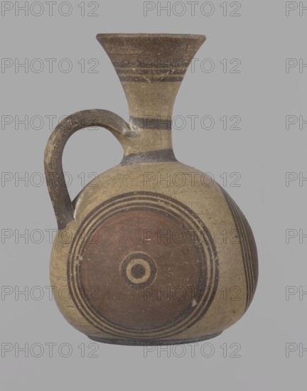 Cypriot jug, Cypriot pottery, Cypriot Bichrome Ware III. Jug on a low, straight foot. Globular body, flattened at the bottom. Quite narrow neck, funnel-shaped spout. Slightly flattened handle attached to the base of the neck and the upper part of the body. Painted decoration in stripes and linear patterns, in red-brown and black fading to brown. Near the spout is a wide red band, below it three black lines, the first adjacent to the red band. At the lower part of the neck, above the handle attachment, a narrow black band; another narrow black band at the base of the neck. The belly is decorated with three identical shooting target motifs on the front and sides: a wide red circular band with a black dot and a circle in the center, surrounded on the outside by four black-brown circles, the first adjacent to the red band. The handle is unpainted on the vessel side, black on the top, with light horizontal lines. The foot on the side is black. On the bottom, two concentric circles with a ce...