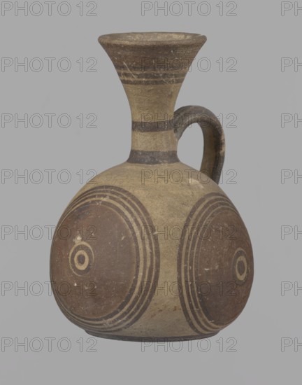 Cypriot jug, Jug on a low, straight foot. Globular body, flattened at the bottom. Quite narrow neck, funnel-shaped spout. Slightly flattened handle attached to the base of the neck and the upper part of the body. Painted decoration in stripes and linear patterns, in red-brown and black fading to brown. Near the spout is a wide red band, below it three black lines, the first adjacent to the red band. At the lower part of the neck, above the handle attachment, a narrow black band; another narrow black band at the base of the neck. The belly is decorated with three identical shooting target motifs on the front and sides: a wide red circular band with a black dot and a circle in the center, surrounded by four black-brown circles, the first adjacent to the red band. The handle is unpainted on the vessel side, black on the top, with light horizontal lines. The foot on the side is black. On the bottom, two concentric circles with a center marked with a dot. Good condition. Cypriot, Cypriot Bi...