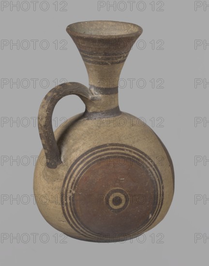 Cypriot jug, Jug on a low, straight foot. Globular body, flattened at the bottom. Quite narrow neck, funnel-shaped spout. Slightly flattened handle attached to the base of the neck and the upper part of the body. Painted decoration in stripes and linear patterns, in red-brown and black fading to brown. Near the spout is a wide red band, below it three black lines, the first adjacent to the red band. At the lower part of the neck, above the handle attachment, a narrow black band; another narrow black band at the base of the neck. The belly is decorated with three identical shooting target motifs on the front and sides: a wide red circular band with a black dot and a circle in the center, surrounded on the outside by four black-brown circles, the first adjacent to the red band. The handle is unpainted on the vessel side, black on top, with light horizontal lines. The foot on the side is black. On the bottom, two concentric circles with a center marked with a dot. Good condition. Cypriot ...