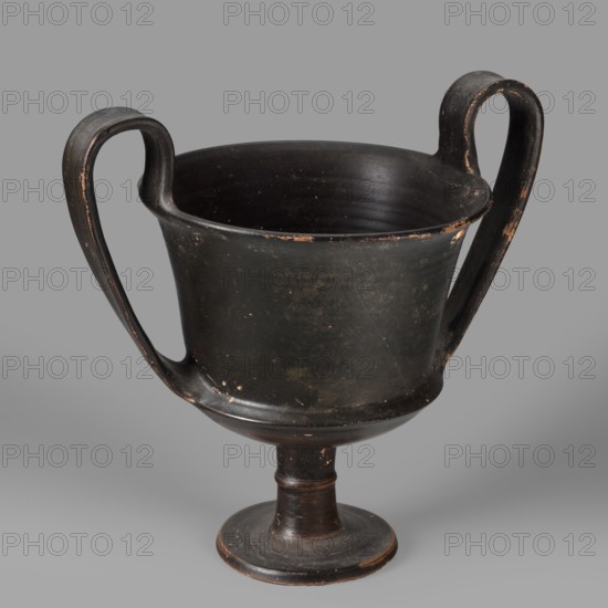 Kantharos, unknown Boeotian workshop. Physical description: Thin-walled kantharos with straight, slightly flared rim edges; a deep cylindrical bowl with an outer edge. The bowl is set on a high leg with a torus in the middle and a flat foot. Band-like, wide handles with slightly raised edges, protruding above the level of the rim. The outer surface of the foot is slightly concave. Decoration: Vessel entirely covered with black varnish. Technology: Matte varnish of good quality, evenly distributed. Traces of turning visible under the varnish. Notes: Numerous secondary losses visible on the surface. Attributed to the 2nd half of the 5th century BC, Boeotia. Kantharos; weight 0.552 kg., A two-handled pedestal vessel with a rounded bowl and flaring rim sits against a neutral background; the body, handles, and foot are primarily black with worn areas revealing brownish and tan underlayers, and there are a few small spots of lighter wear across the surface; the handles curve upward from the ...