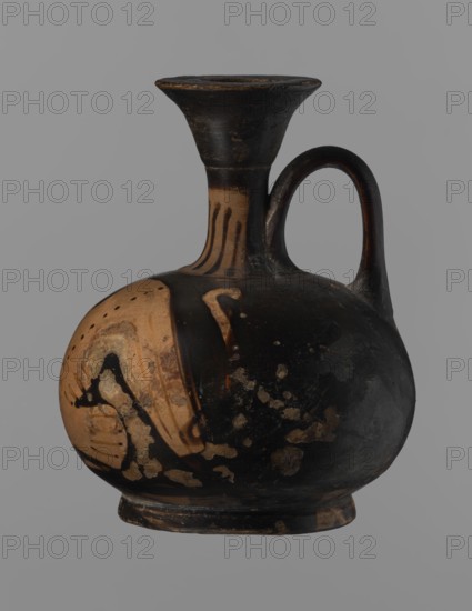 Aryballus lekythos, Ancient Greece, antiquity; bird iconography; ceramics; red-figure pottery; Greek pottery; ancient vessels; lekythos; Southern Italy. Reddish-brown clay with black, slightly shiny, well-covering varnish, also on underside of foot and inside handles. Low echinus-shaped foot, spherical, slightly flattened body. Low, narrow neck with funnel-shaped rim with a thickened edge. Body decoration: bird in left profile with beak raised. 4th century BC Southern Italy. Vessel aryballus lekythos. Height 9.3 cm; width 7.6 cm., A rounded ceramic vessel with a narrow neck, flared rim, and a single curved handle, shown against a plain gray background; the vessel's surface shows a dominant black color with areas of tan and beige where the black finish is worn or intentionally left unpainted, and there are scattered lighter patches and irregular spots on the body; the neck features vertical tan stripes, and the rim and base show worn black with tan edges.