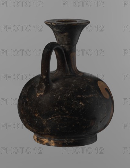 Aryballus lekythos, Reddish-brown clay with black, slightly shiny, well-covering varnish, including underside of foot and inside handles. Low echinus-shaped foot, spherical slightly flattened body; low narrow neck with funnel-shaped rim with thickened lip. Body decorated with a bird in left profile with beak raised. Ceramics, red-figure pottery; Greek pottery, ancient vessel lekythos / aryballus. Southern Italy, 4th century BC. Height 9.3 cm, width 7.6 cm., A rounded, bulbous ceramic vessel with a single curved handle connecting the body to a tall, flared neck and rim; the surface shows worn, chipped areas and a mottled texture. The vessel is predominantly black with patches of brown and tan where the surface has worn away, and there is a small area of lighter brown on one side of the body. The rim and base show visible wear with exposed tan and brown tones, and the handle has small worn spots revealing lighter brown beneath the black. The background is a uniform gray.
