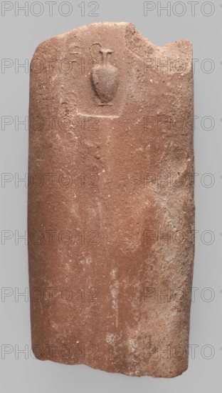 Fragment of a transport amphora vice with a rectangular stamp containing an inscription in two rows and, between them, a depiction of an oinochoe in a horizontal position; incomplete Greek inscription; end of the 4th–beginning of the 3rd century BC; found at Myrmekion; origin Thasos; stamp; height 5.1 cm, width 10.8 cm., A worn, rectangular stone slab with rounded top edges in reddish-brown tones, showing patches of lighter beige and dusty white discoloration and surface roughness; near the upper center there is a shallow, carved outline of a small vessel with a narrow neck and curved handle, the carved area appearing slightly darker and smoother than the surrounding texture; the slab’s right edge has an irregular chipped section and a band of rougher, paler surface, while the left edge is smoother and gently curved; overall the surface displays small pitted marks and speckled lighter spots against the predominant reddish-brown color.