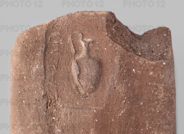 Fragment of a transport amphora vice with a stamp depicting an oinochoe and an incomplete Greek inscription. Fragment of an amphora vice with a rectangular stamp containing an inscription in two rows and between them a depiction of an oinochoe in a horizontal position. End of the 4th–beginning of the 3rd century BC. Found at Myrmekion; origin Thasos. Stamp; vice. Height 5.1 cm, width 10.8 cm., A weathered stone fragment in shades of brown and reddish-brown with a rough, pitted surface and small pale flecks scattered across it, featuring a shallow carved relief of an amphora-like vessel with a rounded body and two handles, a curved ridge along the top edge, and worn irregular edges that show chips and breaks.