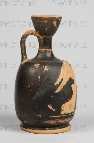 Aryballus lekythos, decoration: on the belly a winged Nike in right profile, dressed in a long chiton, holding a box with both hands above a basket standing on the ground; ca. 460–450 BC; Attica place of manufacture; lekythos vessel; height 13.3 cm, width 8 cm, rim diameter 4 cm, foot diameter 6.4 cm., An upright ceramic vessel with a rounded body, narrow neck, flared rim, and a curved handle attached from neck to shoulder; the surface shows a worn black slip with areas of exposed tan clay where the black has chipped away, creating irregular patches and scratches; the rim, handle edges, shoulder, and base reveal the tan color more prominently; the overall background is a pale neutral tone.