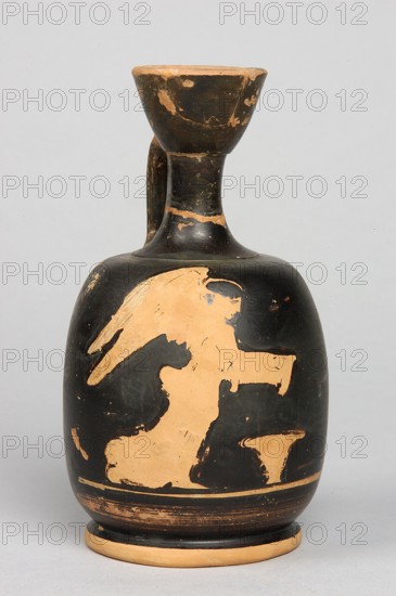 Aryballus lekythos, decoration: on the belly a winged Nike in right profile, dressed in a long chiton, holding with both hands a box with a basket beneath on the ground; ca. 460–450 BC, Attica; lekythos vessel; height 13.3 cm, width 8 cm, rim diameter 4 cm, foot diameter 6.4 cm., A rounded ceramic vessel with a narrow neck and a flared rim sits upright against a plain light-gray background; the vessel is primarily black with areas of worn beige where the surface has chipped or been painted, showing a beige figurative shape on the front that resembles a winged figure in profile, along with additional beige accents near the base and rim; the neck and handle are black with beige wear marks, and the base shows concentric bands of black and beige.