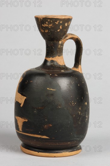 Aryballus lekythos, decoration: on the belly a winged Nike in right profile, dressed in a long chiton, holding with both hands a box with a basket beneath it; ca. 460–450 BC; Attica; lekythos vessel; height 13.3 cm, width 8 cm, rim diameter 4 cm, foot diameter 6.4 cm., A single handled ceramic vessel with a rounded body, a narrow neck, and a flared rim, showing worn surface areas. The vessel is predominantly black with patches of exposed clay in a tan or beige color where the surface has chipped. The handle is curved and also shows black surface with tan worn spots. The background is a plain light gray.