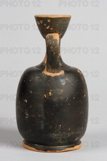Aryballus lekythos, unknown creator; decoration: on the belly a winged Nike in right profile, dressed in a long chiton, holding with both hands a box in front of her, under which a basket stands on the ground; ca. 460–450 BC; Attica place of manufacture; lekythos vessel; height 13.3 cm, width 8 cm, rim diameter 4 cm, foot diameter 6.4 cm., A single ceramic vessel with a rounded body and a narrow neck flaring into a small cup-like rim, showing a worn black surface with patches where the underlying clay color shows through; the handle runs from the rim to the shoulder and shares the same black finish with worn areas; the base reveals the natural clay color around its edge; the background is a plain light gray.