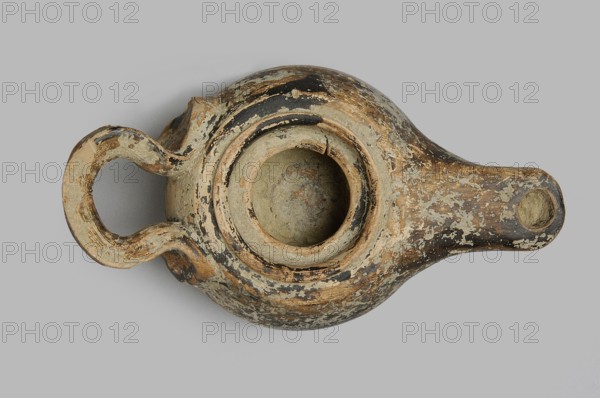 Oil lamp, unknown Attic workshop. Wheel-made lamp with a globular, slightly squat container. Raised, round base, slightly concave unglazed. Downward-sloping arms bounded by a distinct shaft and groove. Large oil opening. Long, flat burner. Ribbon-shaped vice, placed almost horizontally. Black/dark brown firnis on the surface outside the base. 360–300 BC. Attica. Vessel; length 13.3 cm, width 7.2 cm, height 4 cm., An overhead view of an aged oil lamp with a rounded body, a broad circular opening at its center, a curved handle on one side, and a narrow spout on the opposite side; the surface shows peeling layered paint and wear with visible areas of beige, tan, brown, black, and gray, and the background is a uniform light gray.