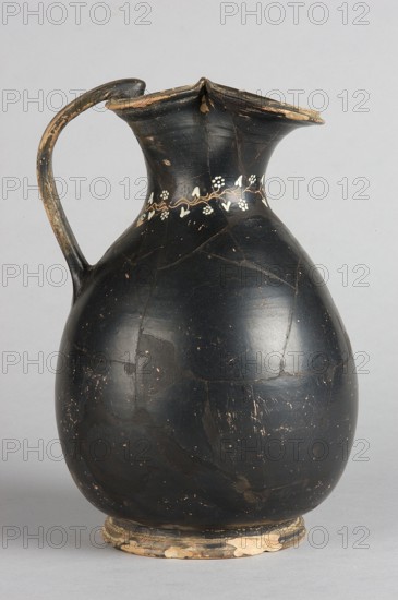 Oinochoe, Gnathia style. Decoration: on the neck an engraved horizontal ivy sprig with white-painted heart-shaped leaves and flowers in the shape of pointed rosettes, and a second one extending downwards from it, placed centrally at the front. 2nd half of the 4th century BC, Apulia. Oinochoe vessel, height 19.5 cm, width 13 cm., A single tall ceramic jug with a rounded body, narrow neck, and flared irregular rim, shown against a plain light gray background; the jug is predominantly black with areas of surface wear revealing lighter brown and beige beneath, a curved handle attached from rim to shoulder with similar wear, a decorative band of small white floral or vine-like motifs encircling the neck, fine hairline cracks and scattered chips across the surface, and a worn beige and dark base that shows flakes of exposed clay.