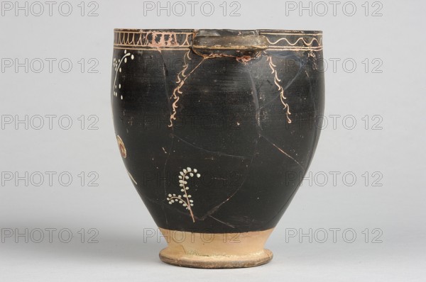 Skyphos with painted decoration. Engraved ornament at the upper edge: an egg-shaped motif and a wavy line, framed by parallel lines. At the handles and in the center, vertical engraved branches with preserved traces of white leaves hang downwards. Side A: a woman shown in left profile with a xylophone in her right hand and a branch in her left. The figure is flanked by small trees on either side. 4th century BC, Apulia, Southern Italy. Skyphos vessel, height 12.1 cm, width 11.1 cm., A dark rounded ceramic vessel with a small rim and a narrow base sits against a pale background, showing a predominantly black surface with areas of worn lighter brown and tan, and decorative pale peach squiggles and small pale dots forming simple botanical motifs; the base and foot are tan and light brown with some greenish residue, and there are visible fine cracks and chipped edges on the rim and body.
