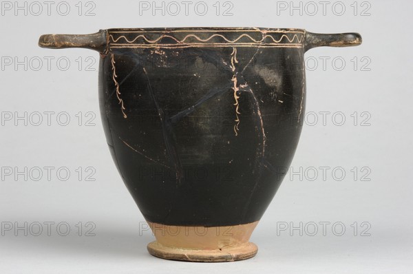 Skyphos, Skyphos with painted decoration. Engraved ornament at the upper edge of the vessel: an egg-shaped structure and a wavy line, framed by parallel lines. At the handles and in the center, vertical, engraved branches with preserved traces of white leaves hang downwards. Side A: a woman shown in left profile with a xylophone in her right hand and a branch in her left. The figure is flanked by small trees on either side. 4th century BC, Apulia, Southern Italy. Skyphos vessel, height 12.1 cm, width 11.1 cm, A single black pottery vessel with two horizontal handles extending from opposite sides, showing worn areas and thin decorative squiggled lines near the rim in a lighter clay color; the lower portion of the vessel and its flared foot reveal exposed terracotta tones, and the surface shows hairline cracks and lighter scuffed patches; the background is a pale gray.