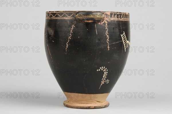 Skyphos with painted decoration. Engraved ornament at the upper edge of the vessel: an egg-shaped structure and a wavy line, framed by parallel lines. At the handles and in the center, vertical, engraved branches with preserved traces of white leaves hang downwards. Side A: a woman shown in left profile with a xylophone in her right hand and a branch in her left. The figure is flanked by small trees on either side. 4th century BC, Apulia, Southern Italy. Skyphos vessel. Height 12.1 cm, width 11.1 cm., A rounded ceramic vessel with a narrow base and flared rim sits against a pale gray background; the body is predominantly black with areas of worn terracotta showing through near the rim and base, and there are decorative pale cream and faded pink linear and dotted motifs on the black surface, including a curving dotted vine-like element and wavy vertical lines, plus a small protruding handle near the top that shows chipped black and exposed terracotta.