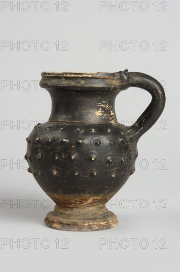 Black-finned oinochoe, decoration: in the upper half of the belly four rows of plastic knobs applied using the barbotine technique, IV waC, Campania, vessel, height 9.8 cm, width 6.7 cm, An aged rounded vessel with a single curved handle displayed against a light gray background; the vessel is predominantly black with worn areas showing beige and brown beneath, a textured body featuring a pattern of raised circular bumps, a flared neck and rim with chipped edges revealing lighter color, and a footed base that shows patches of brown, beige, and gray.