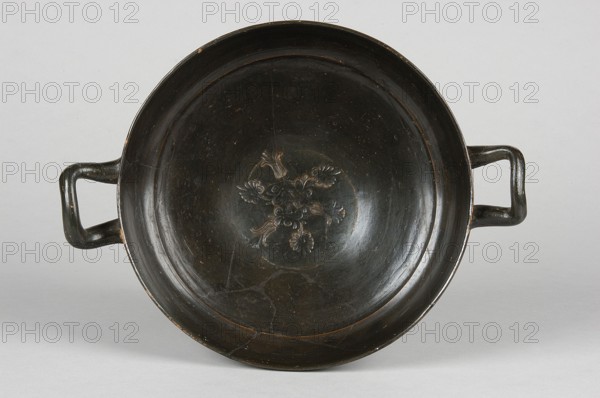 Kyliks, stamped decoration: at the bottom of the bowl, 3 radially arranged palmettes and 3 lotus flowers arranged in pairs, connected by a branch. IV waC, Campania, kylix vessel, A shallow round bowl with two rectangular loop handles lies on a light background; the bowl is dark brown to black with a slightly glossy surface and shows a decorative raised floral motif in the center in a similar dark tone with some lighter brown highlights, and there are visible scuffs and worn edges revealing lighter brown beneath the dark finish.