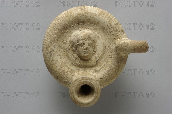 Guttus, top decorated with a plastic medallion with the image of a woman's head shown three-quarters to the right, fluted arms, III waC, Apulia, guttus vessel, height 7.4 cm, width 8.0 cm, A circular, worn clay object in beige and tan tones with rough, textured surfaces and patches of lighter and darker beige sits against a smooth gray background; the object has a raised central medallion featuring a worn human face with curly hair, a round rim with irregular edges, a short spout extending from one side, and a wider tubular opening at the lower edge, all showing weathered, grainy texture and subtle variations of cream and sandy brown.
