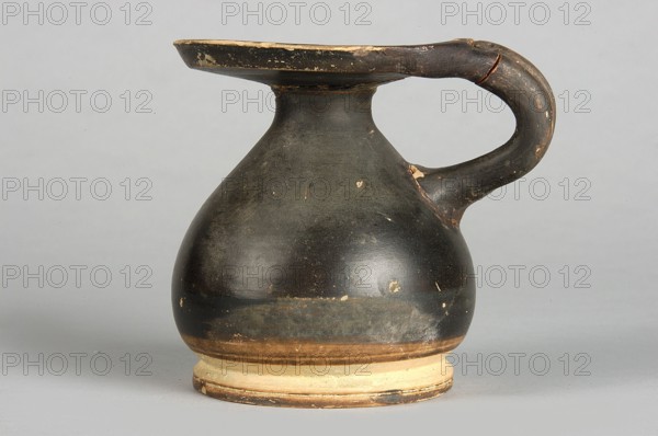 Black-finned alabastron, wide disc-shaped foot, circular body with sloping shoulders, narrow neck, wide horizontal rim and vertical bipartite handle; end of 4th–3rd century BC, Southern Italy; alabastron vessel; height 7.5 cm, width 6.7 cm., A rounded ceramic jug with a single looped handle and a wide flat rim, showing a worn dark surface with lighter areas where the finish is chipped. The jug displays colors including dark blackish-brown on the body and handle, muted tan and beige on the base and rim edges, and small spots of pale cream where the surface is worn. The background is a smooth light gray.