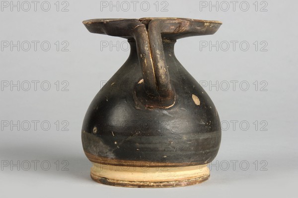 Black-finned alabastron, wide disc-shaped foot, circular body with sloping shoulders, narrow neck, wide horizontal rim and vertical bipartite handle; end of 4th–3rd century BC, Southern Italy; alabastron vessel; height 7.5 cm, width 6.7 cm., An old rounded vessel with a flared rim and a single looped handle is centered against a plain light gray background, the vessel showing a worn dark brown to black surface with patches where the underlying lighter clay is exposed, a lighter beige band around the base, and visible scratches and chips revealing tan and ochre tones.
