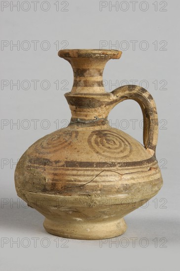 Jug, painted decoration: in the widest part of the body, on the neck and handle - horizontal lines of various widths; on the shoulders - 4 groups of 4 concentric circles; on the spout - concentric lines and small semicircles; 850–750 BC; Cypriot, Geometric Period III, Cyprus; vessel; height 7.5 cm, width 6.8 cm, A single ceramic jug with a rounded body, narrow neck, flared rim, and a curved handle is shown against a plain light gray background. The jug displays concentric circular patterns and horizontal bands in darker earth tones on a base of light beige clay; the colors visible include beige, tan, brown, and hints of ochre. The surface shows areas of wear and small chips, with a visible crack and some rough, textured patches where the clay appears abraded or repaired. The handle and neck have alternating lighter and darker horizontal stripes, and the rim has worn darker edges. The jug rests on a short footed base that is slightly lighter in color than the body.