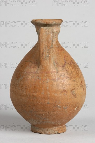Pelike, Decoration painted on the firnis: between the handles on both sides, a wavy linear tendril with volute-like curled side shoots; above it, a wavy line twist?. At the height of the base of the handles, two horizontal parallel lines. Date: 332–30 BC Hellenistic period., Vessel; height 12.5 cm, width 9.8 cm., A rounded ceramic vessel with a narrow neck and a single vertical handle, standing on a small circular foot, with an overall warm terracotta color and areas of lighter beige and grayish weathering; the surface shows a few darker reddish-brown patches and some pale greenish spots, with a slightly rough, worn texture visible across the body and handle, set against a neutral light gray background.