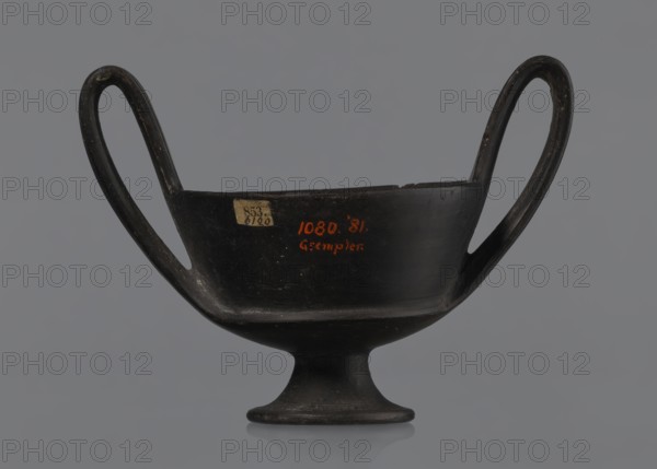 Kantharos, Kantharos black-varnished on a cup-shaped foot, vessel; 1st half of the 6th century BC, Etruria Italy; function: goblet for banquets; provenance: repossession Bytom., A dark, two-handled vessel with a shallow bowl and a flared foot sits against a neutral gray background, the vessel appearing predominantly black with subtle variations in dark gray where light reflects, and small areas of worn lighter gray on the handles and rim; the handles form elongated loops rising above the bowl on each side, and the interior and exterior surfaces show a slightly glossy finish with faint scuff marks and texture.