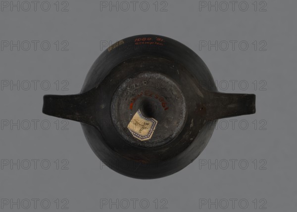 Kantharos, Etruria Italy, repossession Bytom, vessels, goblets, banquets, Italy, antiquity, kantharos black-varnished on a cup-shaped foot, 1st half of the 6th century BC, Etruria, vessel, A top-down view of a dark brown rounded vessel with a central circular opening and a short neck, flanked by a horizontal handle-like extension on either side; the vessel surface shows areas of lighter brown wear and a few small scuffs, with a small pale sticker-like piece affixed near the neck; the background is a flat muted gray.