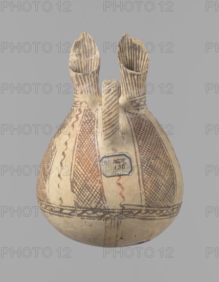 Jug, unknown Cypriot workshop. White painted ware jug with a spherical bottom, a bulging pear-shaped body transitioning in the upper part into two tubular spouts with protruding walls. Between them, a ring-shaped vise protrudes from the body. The vessel is covered with white paint and decorated with brownish-red paint. A zigzag ornament runs around the body for 1/3 of the vessel's height, flanked on each side by two horizontal lines. Below are four bands of lines converging at the bottom. The upper part of the body is decorated with four stripes filled with diagonal, intersecting lines. The stripes are separated by lines that wind serpentine. The vise is decorated with diagonal lines at the top and brown paint on the sides. At the base of the rims, a zigzag ornament is framed by two lines on either side; above it, vertical lines surround the rims both inside and outside. Bronze Age, 1900–1650 BC, Cyprus, vessel. Height 15 cm, width 13 cm., A rounded ceramic vessel with a bulbous body a...