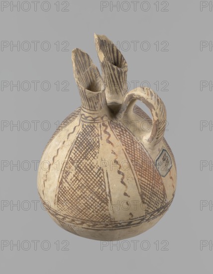 Jug, unknown Cypriot workshop. White painted ware jug with a spherical bottom and bulging, pear-shaped body transitioning in the upper part into two tubular spouts with protruding walls. Between them, a ring-shaped vise protrudes from the body. The vessel is covered with white paint and decorated with brownish-red paint. A zigzag ornament runs around the body for 1/3 of the vessel's height, flanked on each side by two horizontal lines. Below are four bands of lines converging at the bottom. The upper part of the body is decorated with four stripes filled with diagonal, intersecting lines, the stripes separated by serpentine winding lines. The vise is decorated with diagonal lines at the top and brown paint on the sides. At the base of the rims, a zigzag ornament is framed by two lines on either side; above it, vertical lines surround the rims both inside and outside. Bronze Age, 1900–1650 BC, Cyprus. Vessel; height 15 cm, width 13 cm., A rounded ceramic vessel with a handle and three u...