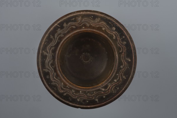 Black-finished bowl, 4th century BC, Southern Italy; vessel bowl; height 6.0 cm, width 11.5 cm., A round shallow bowl viewed from above with a wide rim and a recessed center, showing a dark brown to black surface with lighter brown and reddish accents; the rim features a repeating wavy decorative pattern in lighter beige and reddish tones against the darker background, and there are small raised dots and worn spots around the rim; the central recessed area is darker and smoother with subtle circular marks, and the background behind the object is a uniform soft gray.