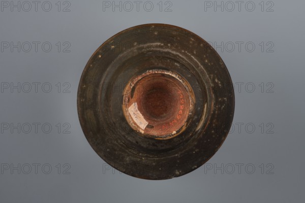 Black-finished bowl, 4th century BC, Southern Italy; vessel bowl; height 6.0 cm, width 11.5 cm., A top-down view of a circular object resembling a bowl or vessel with a wide rim and a recessed center, showing a worn, textured surface in dark brown and black tones with patches of lighter brown and ochre; the central interior has reddish-brown and terracotta hues with a few areas of chipped lighter beige and pale tan; the background is a smooth gray-blue.