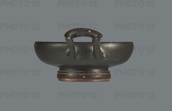 Black-finished goblet, 4th century BC, Campania; vessel goblet; height 5.0 cm, width 12.5 cm., A dark, shallow bowl-like object with a rounded rim and a raised circular foot, shown from the side against a uniform gray background. The object is predominantly black with areas of worn brownish-orange showing through along the foot and small worn spots on the rim and body. A curved handle rises from the interior and connects to the rim with two thick supports, all sharing the same black surface with small scuffs and patches of lighter wear. The surface has a subtle sheen and visible wear marks, and the overall shape is symmetrical with smooth, rounded contours.