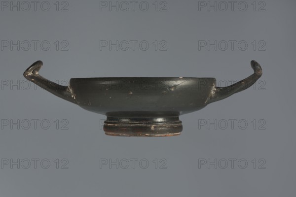 Black-finished goblet, 4th century BC, Campania; vessel goblet; height 5.0 cm, width 12.5 cm., A shallow, dark bowl with a glossy black surface and a rounded foot sits centered against a neutral gray background, showing wear with small chips and lighter scratches; it has two elongated handles that curve upward and outward, each handle ending in a rounded tip, and there are subtle brownish areas near the foot and on the worn spots.