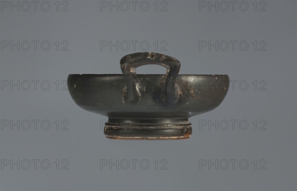 Black-finished goblet, 4th century BC, Campania; vessel goblet; height 5.0 cm, width 12.5 cm., A dark, shallow bowl-like object with a rounded base and a short pedestal, shown against a solid gray background; the object is predominantly black with areas of worn brown and subtle reddish-brown showing through, and it has a curved handle rising from the rim with visible scuffs and patches of lighter wear on its surface.