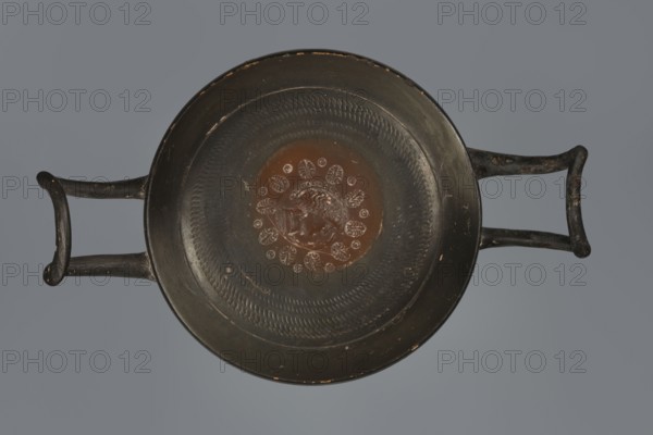 Black-finished goblet, 4th century BC, Campania; vessel goblet; height 5.0 cm, width 12.5 cm., A top-down view of a shallow round dish with two opposing handles, showing a predominantly dark surface with a central circular area in a reddish-brown hue. The rim and handles are dark, almost black, with small worn spots revealing lighter tones. Around the central reddish-brown area is a pattern of raised petal-like shapes and concentric textured bands in dark and slightly lighter shades. The background behind the dish is a muted gray.
