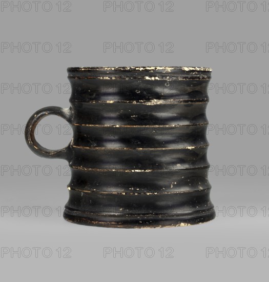 Dice cup. Small cup with a flat slightly concave bottom, slightly tapered walls, distinct rim and foot. Outer surface decorated with seven wide ribs; bottom has concentric decoration. Entire object covered with black varnish shiny but matte in places; traces of turning visible under varnish. Spout slightly chipped; surface shows dirt and secondary losses; varnish worn on rib ridges. Clay color 5 YR 6/6 reddish yellow; varnish color 2.5 YR 2.5/1 black. Dice game. 5th century BC, Attica. Vessel cup. Height 8.3 cm; width 10.6 cm; depth 8.3 cm., A worn black ceramic mug with a round handle on one side, featuring a ribbed, undulating surface with a few chips and worn areas revealing lighter material beneath; the background is a flat gray.