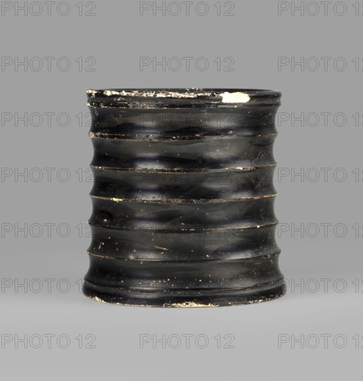 Dice cup. Small cup with a flat slightly concave bottom; slightly tapered walls and distinct rim. Outer surface decorated with seven wide ribs; distinct foot. Entire object covered with black varnish shiny but in places matte; good quality except for a small fragment on the inner surface and the outer surface of the bottom, which has concentric decoration. Traces of turning visible under the varnish. Spout slightly chipped in one place; surface shows dirt and secondary losses; varnish worn on rib ridges. Clay color 5 YR 6/6 reddish yellow; varnish color 2.5 YR 2.5/1 black. 5th century BC, Attica. Vessel cup. Height 8.3 cm; width 10.6 cm; depth 8.3 cm., A cylindrical object with a ribbed, wavy surface sits centered against a neutral background; the object is predominantly black with worn areas showing light beige or off-white flecks and patches, its surface has a slightly glossy finish with subtle lighter black and gray smudges, and the background appears gray.