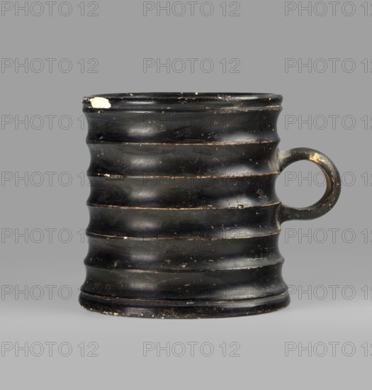 Dice cup, Goluchów Greater Poland Voivodeship. Greek ceramic dice game cup, 5th century BC, Attica. Vessel with small flat slightly concave bottom, slightly tapered walls, distinct rim and foot; outer surface decorated with seven wide ribs. Object entirely covered with black varnish shiny but in places matte; traces of turning visible under varnish, except for a small fragment on the inner surface and the outer surface of the bottom, which has a concentric decoration. Condition: spout slightly chipped in one place; surface shows dirt and secondary losses; varnish worn on rib ridges. Clay color 5 YR 6/6 reddish yellow. Varnish color 2.5 YR 2.5/1 black. Dimensions: height 8.3 cm, width 10.6 cm, depth 8.3 cm., A dark, ribbed cylindrical cup with a rounded handle on one side, showing a few scuffed and worn areas where a lighter color shows through; the cup appears glossy black overall with subtle variations in sheen and small patches of brownish wear, set against a smooth gray background.