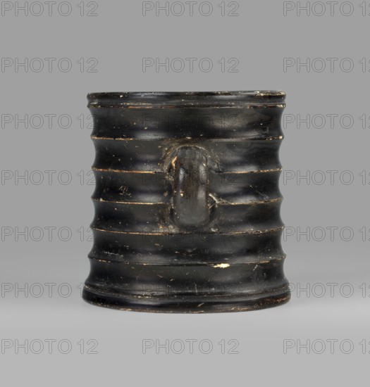 Dice cup. Small cup with flat slightly concave bottom; slightly tapered walls; distinct rim and distinct foot. Outer surface decorated with seven wide ribs. Entire object covered with black varnish shiny but in places matte, except for a small fragment on the inner surface and the outer surface of the bottom, which has a concentric decoration. Traces of turning visible under the varnish. Spout slightly chipped; surface shows dirt and secondary losses; varnish worn on rib ridges. Clay color: 5 YR 6/6 reddish yellow. Varnish color: 2.5 YR 2.5/1 black. 5th century BC, Attica. Vessel cup. Height 8.3 cm; width 10.6 cm; depth 8.3 cm., A ribbed cylindrical vessel with a single rounded handle projecting from its side, shown against a neutral gray background; the vessel is predominantly black with small areas of worn lighter brown and off-white showing through its surface, and it has a slightly glossy finish with visible scuffs and chips.